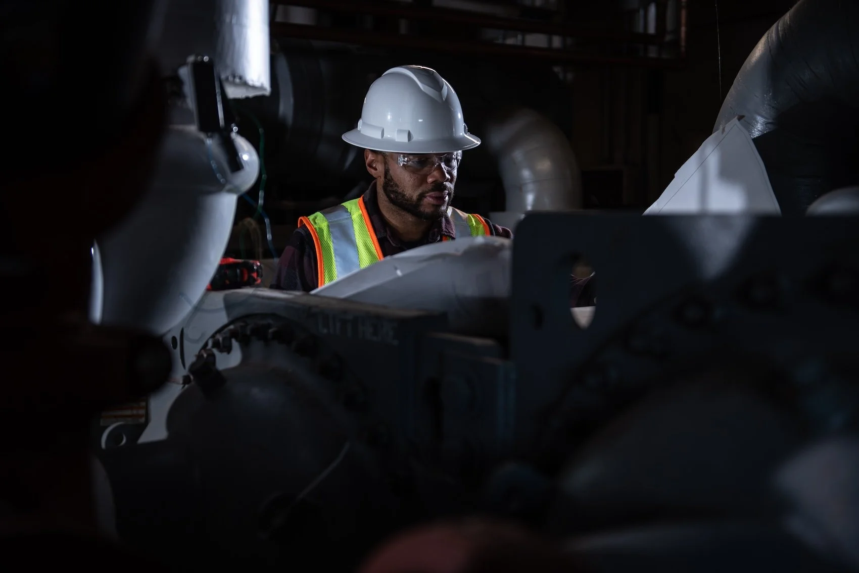 A male worker in a hard hat and safety vest inspecting machinery in a dark industrial setting.