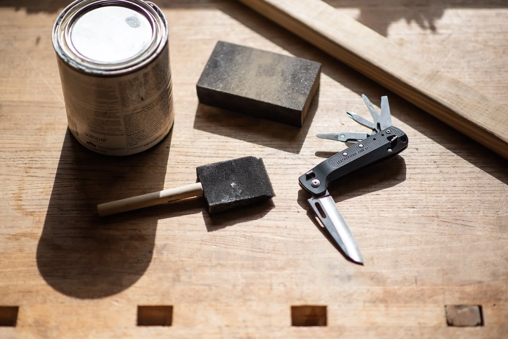 A woodworking workspace with a can of Valspar paint, two sanding blocks, a small hammer, and a multi-tool with screwdrivers on a wooden surface.
