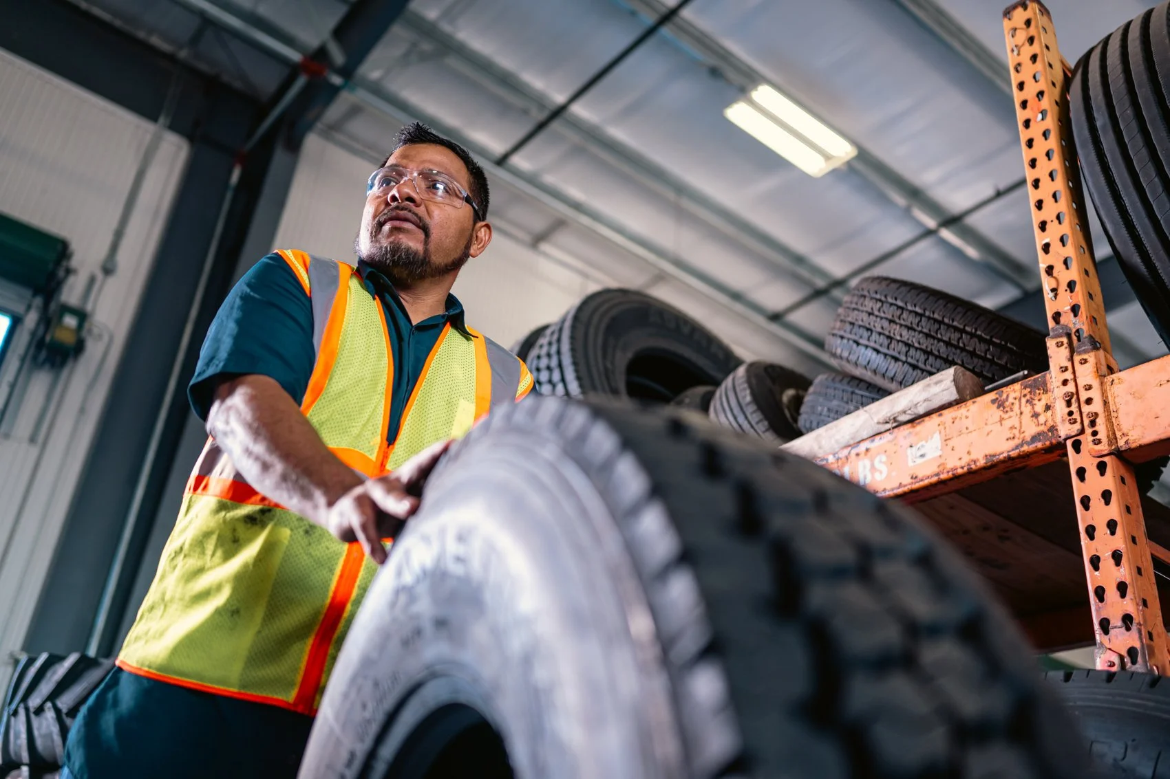 A man wearing a safety vest examines a tire in a warehouse with shelves of tires visible in the background.