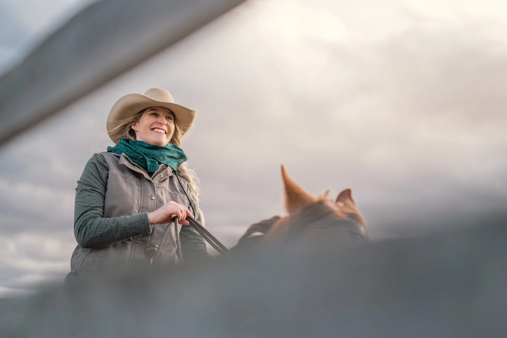 A woman wearing a cowboy hat, a green scarf, and a gray vest, smiling while riding a horse against a cloudy sky.