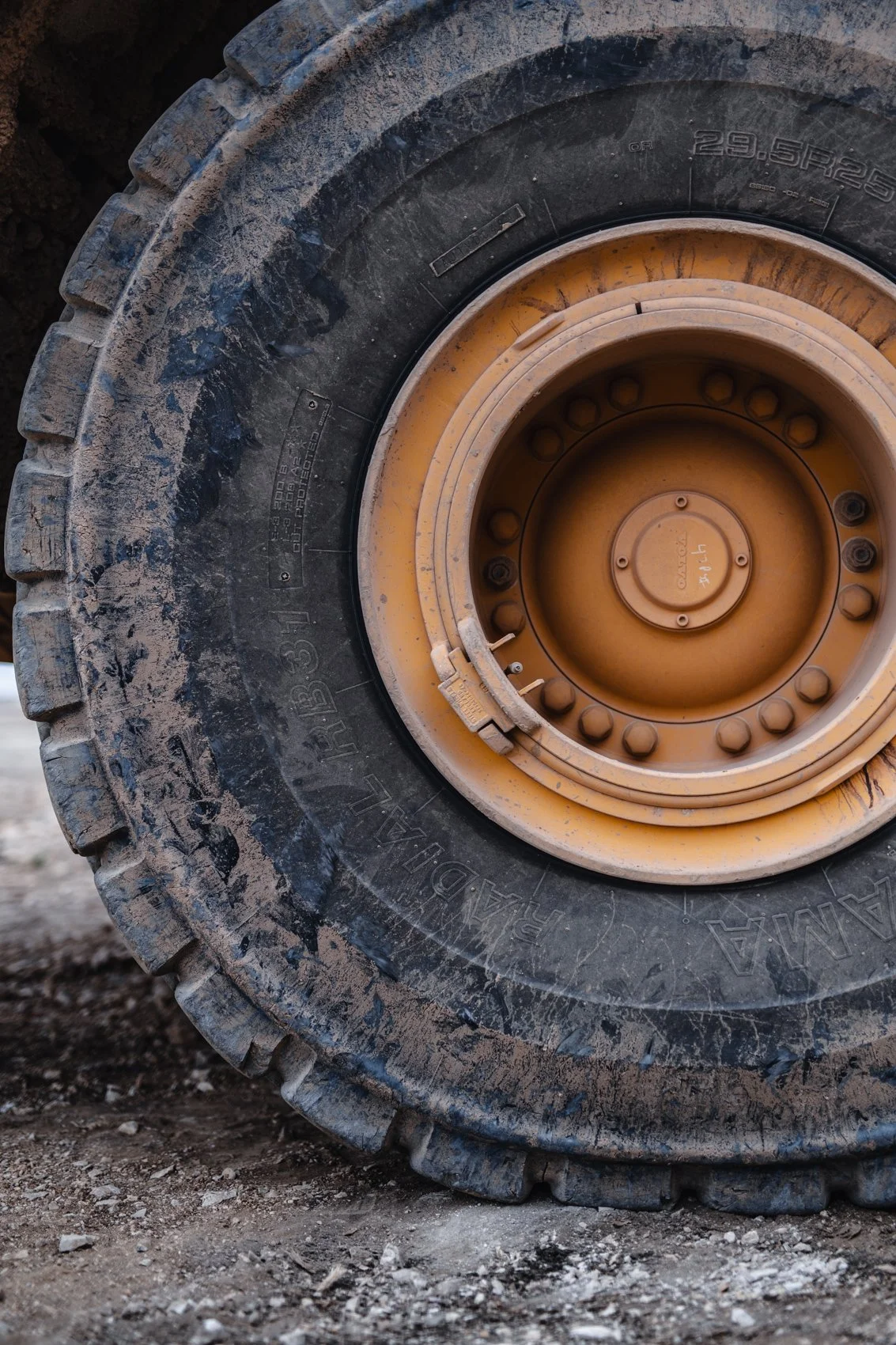 Close-up of a large construction or off-road tire with dirt and mud on the treads, attached to a yellow rim on the ground.