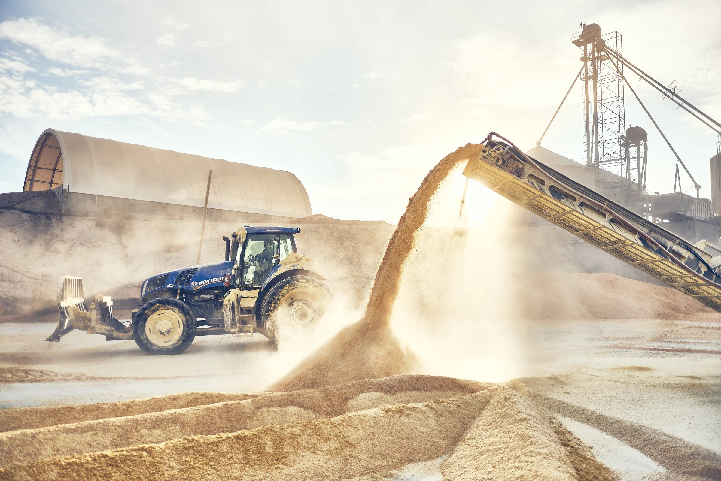 A blue tractor operates on a sandy construction site with a conveyor belt pouring sand into a pile. Large storage structures and a crane are visible in the background under a partly cloudy sky.