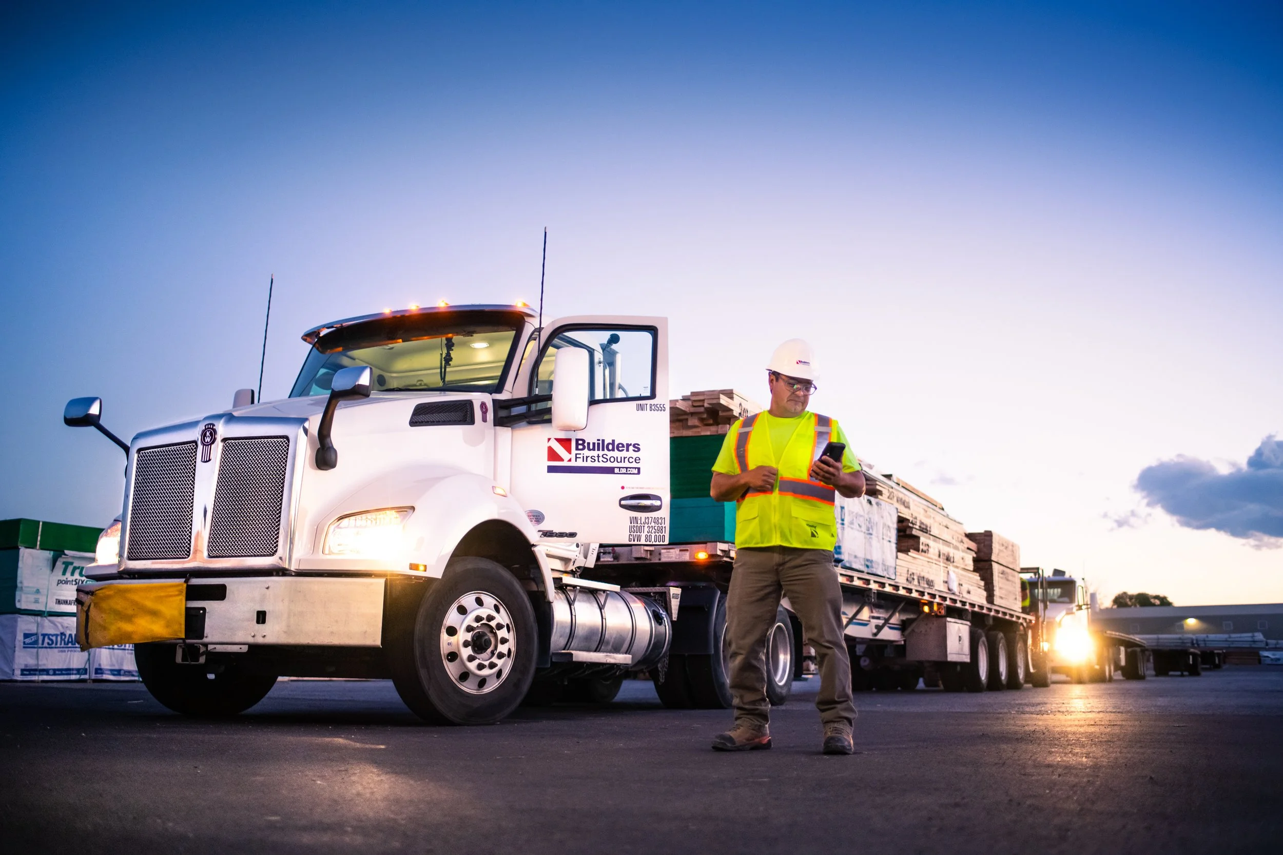 Construction worker in yellow safety vest and white hard hat looking at smartphone next to a white flatbed truck loaded with wooden pallets at dusk