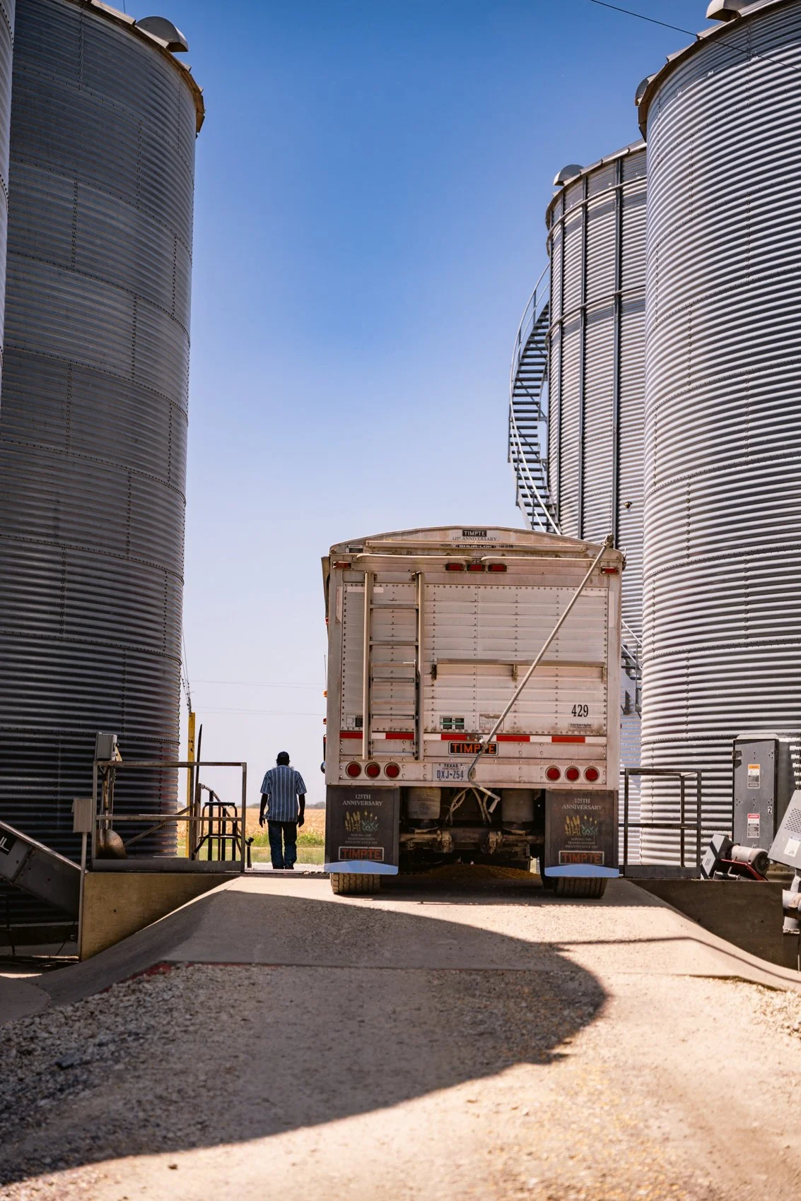 A large white truck parked between two tall, cylindrical metal grain silos on a farm, with a man walking away from the truck towards an open field in the background during daytime.