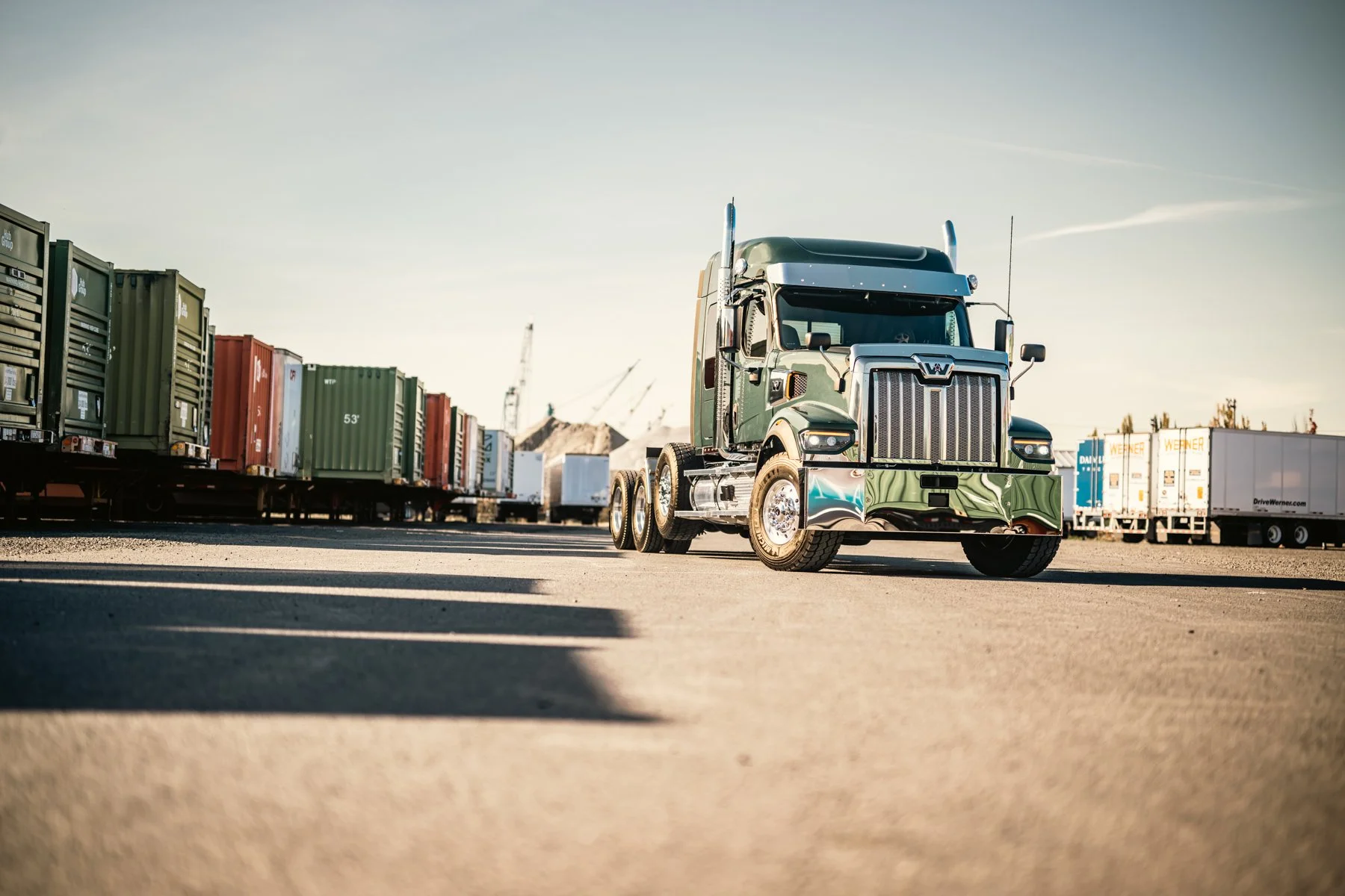 Semi-truck parked in a shipping yard with cargo containers and cranes in the background during daylight.