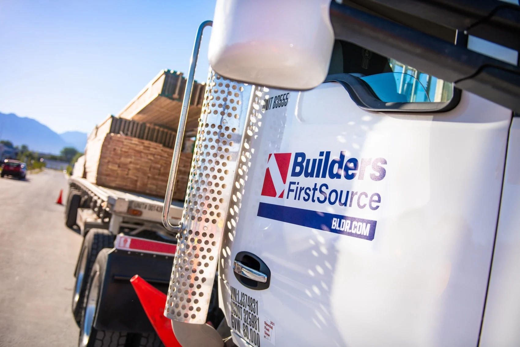Close-up of a construction delivery truck with a "Builders FirstSource" logo, loaded with bricks, on a sunny road with mountains in the background.