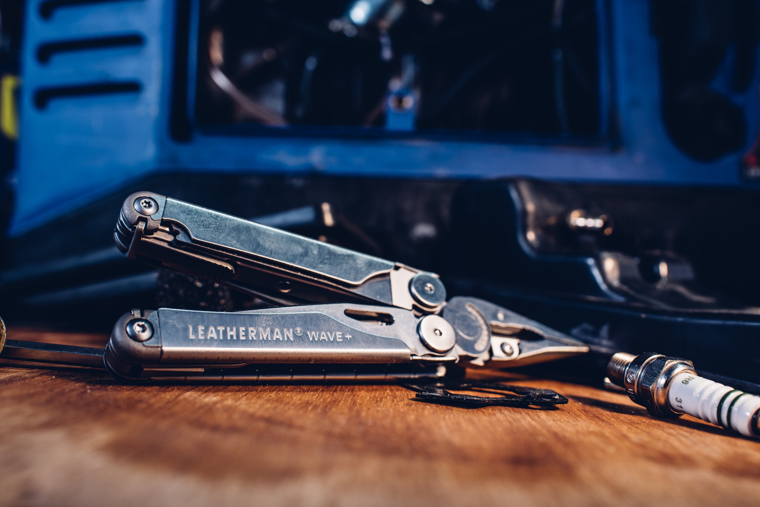 A leatherman multitool labeled 'LEATHERMAN WAVE+' resting on a wooden surface with a spark plug nearby and a toolbox in the background.