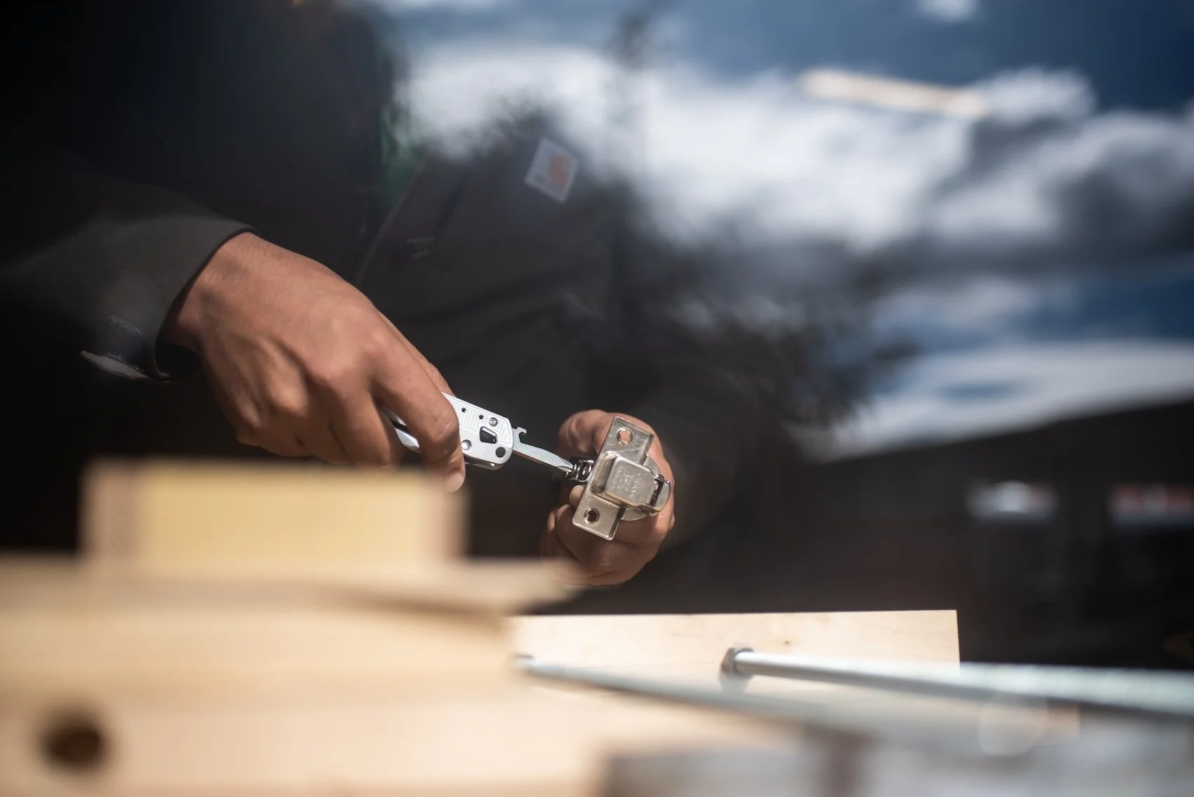 A person using a cordless power drill to fasten a screw into a piece of wood, with a cloudy sky reflected in the window behind them.