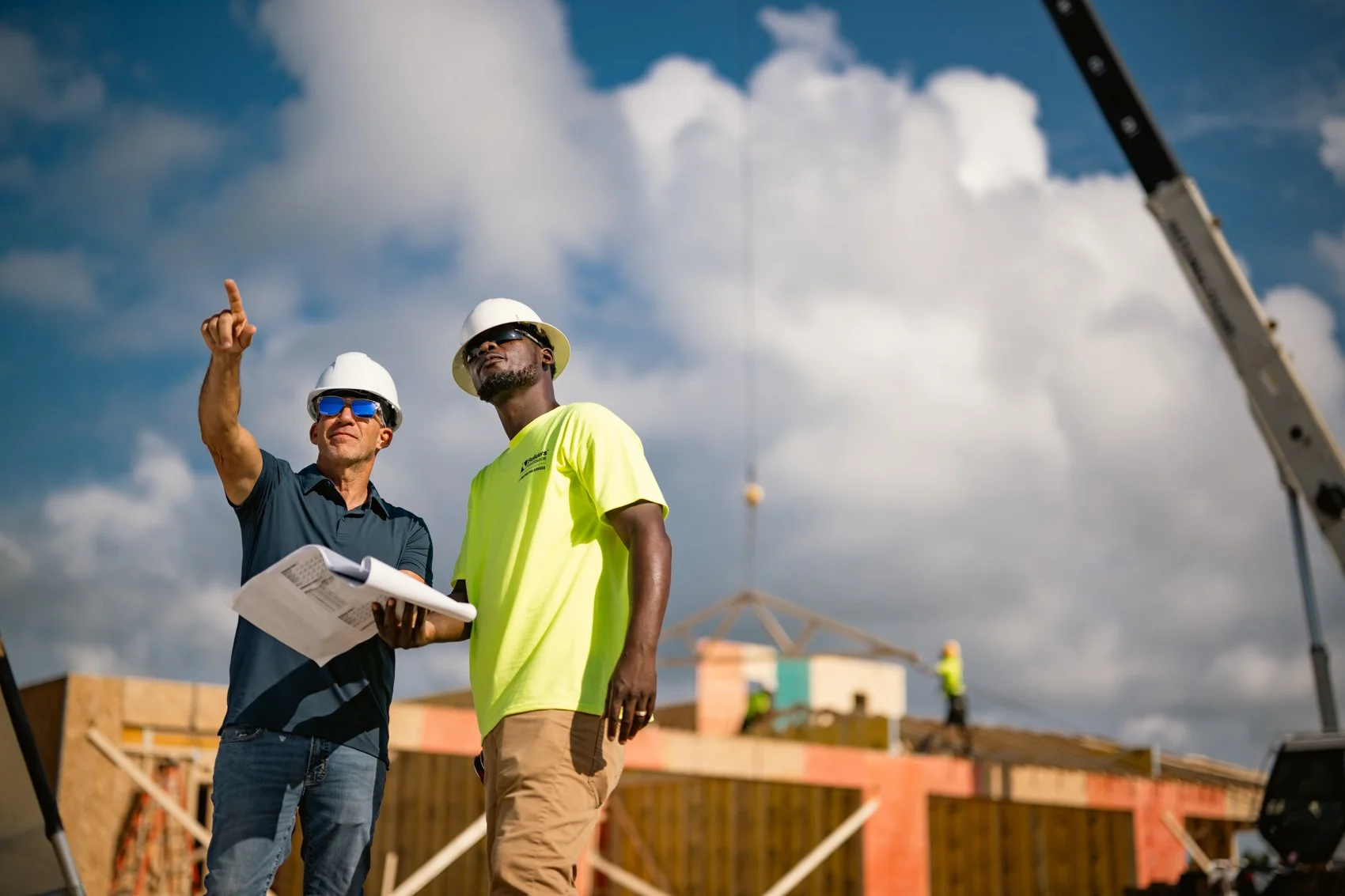 Two construction workers wearing helmets and safety glasses standing on a building site with a crane in the background, discussing plans and looking at the sky.
