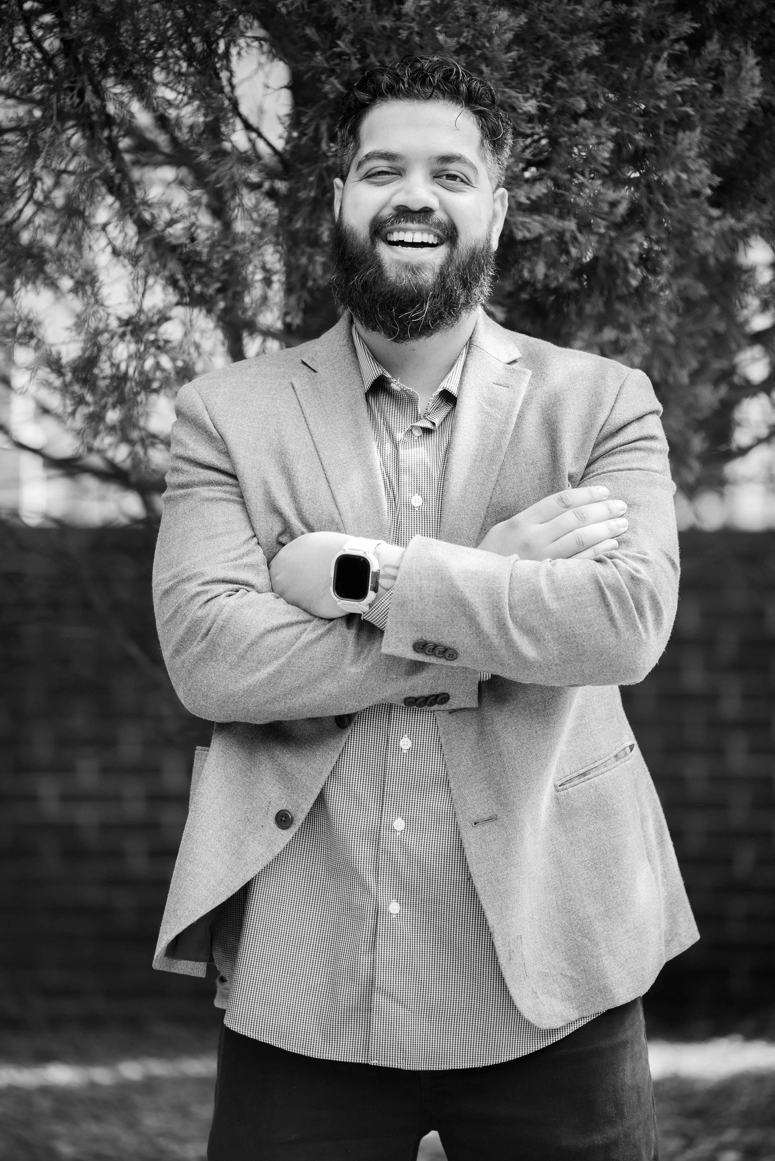 A man with a beard and curly hair, smiling and crossing his arms, wearing a blazer and a smartwatch, standing outdoors in front of a tree.
