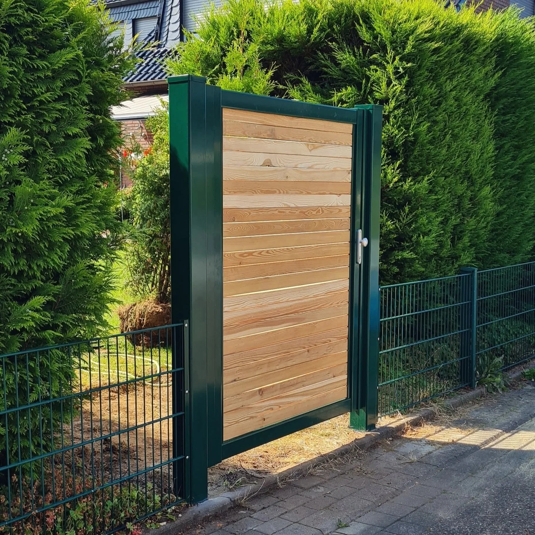 Ein grüner Metallzaun mit einem Torelement aus Holz, das den Zugang zu einem Garten oder Grundstück ermöglicht.
