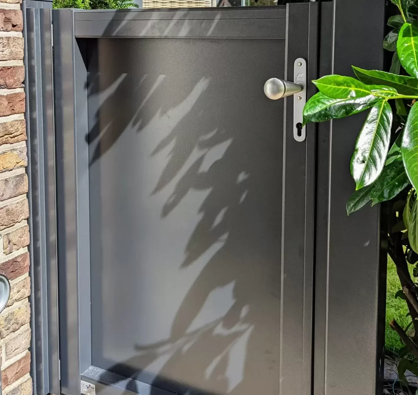Ein grauer Briefkasten mit einem runden Silberknauf vor einer Ziegelmauer und umgeben von grünen Blättern, mit Schatten auf der Tür.