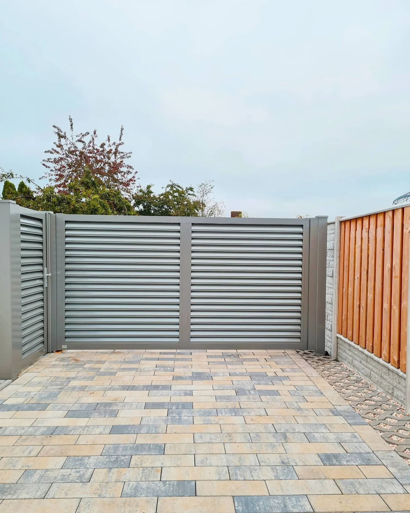 Ein geschlossener Metallzaun mit horizontalen Lamellen, umgeben von einer Terrasse mit Pflastersteinen, neben einer Holzwand und einigen Bäumen im Hintergrund.