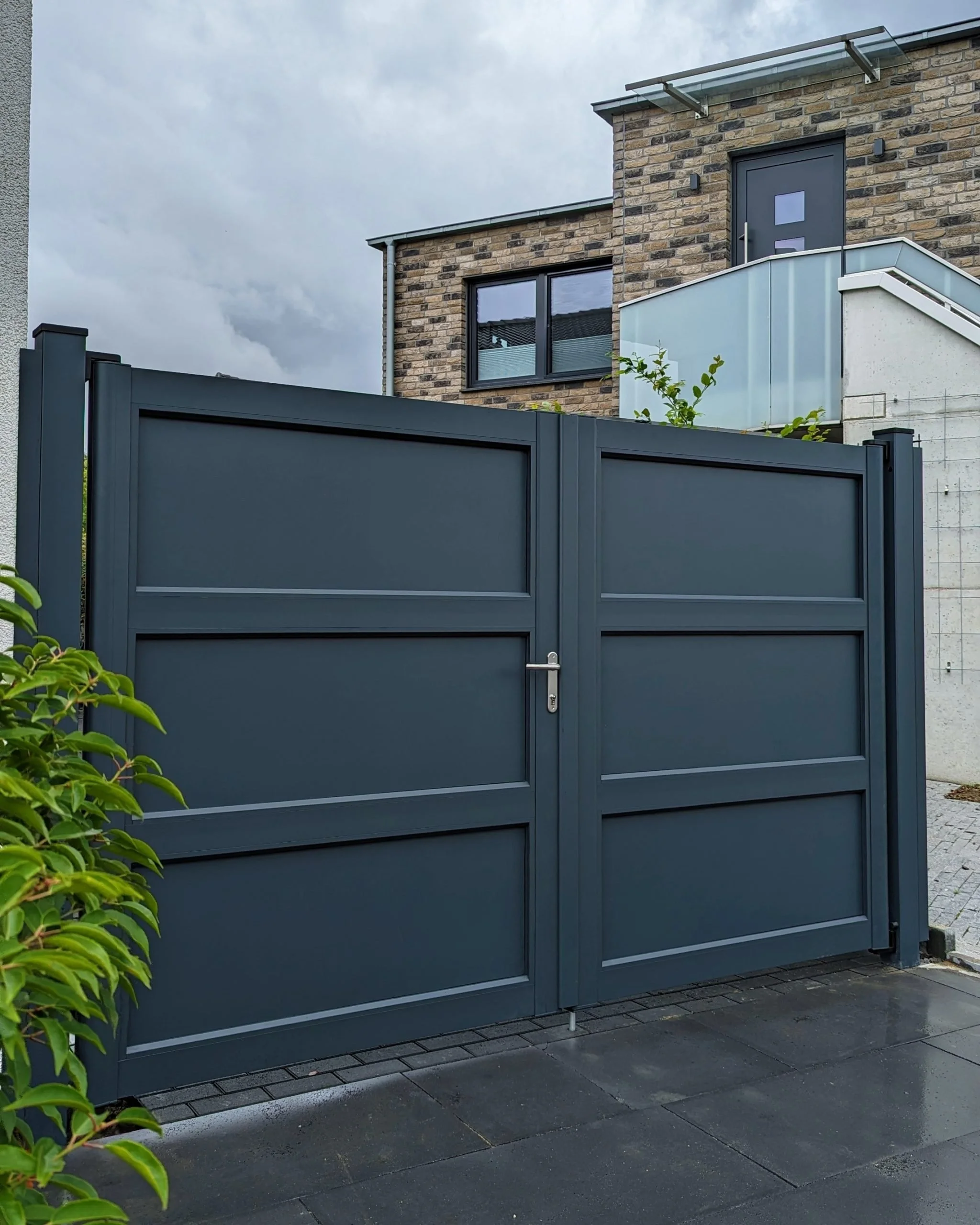 Ein schwarzer Metallzaun vor einem modernen Wohnhaus mit Backstein und einer weißen Balkonbrüstung. Wolkiger Himmel im Hintergrund.