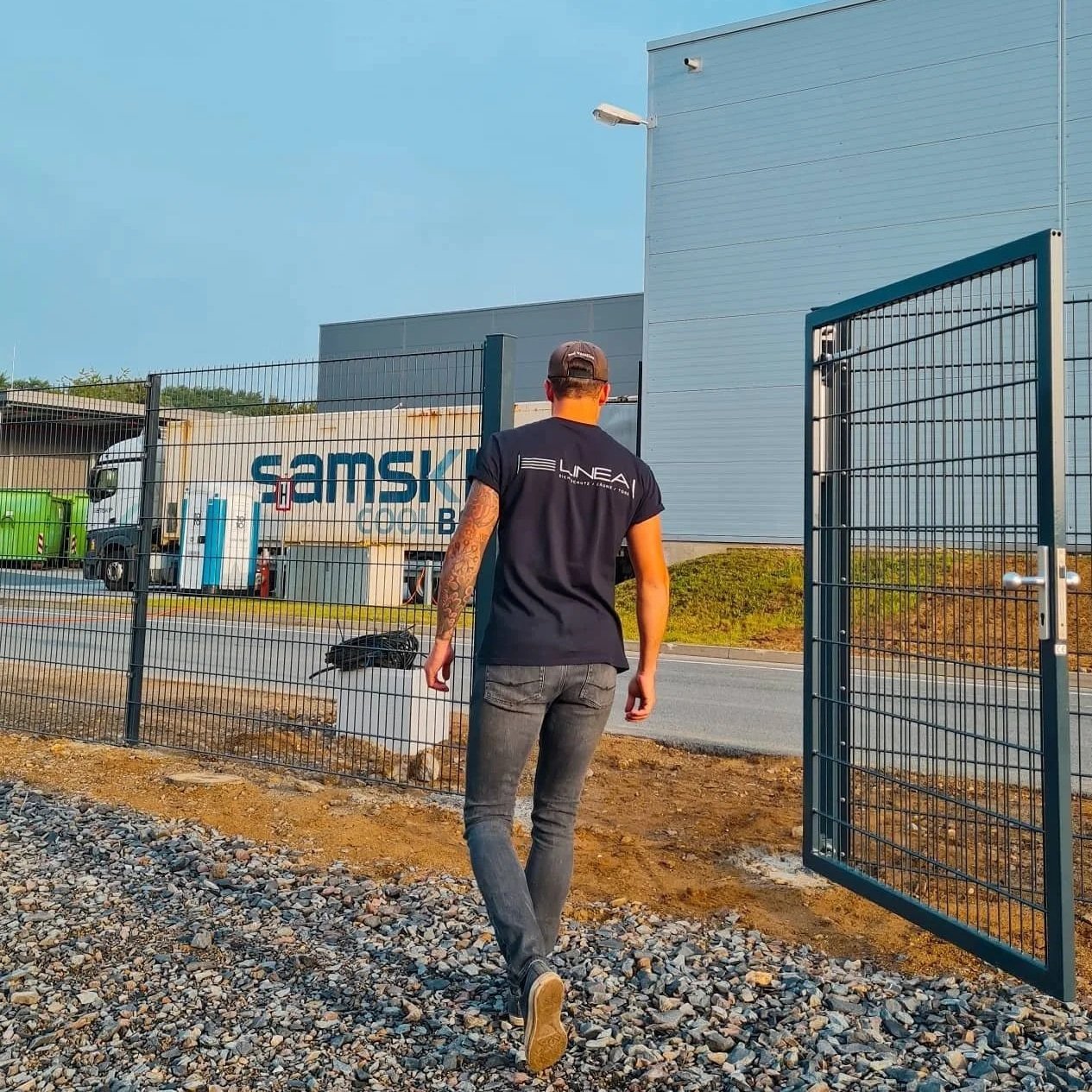 Ein Mann mit Tattoos trägt ein schwarzes T-Shirt und eine Kappe, während er an einem Zaun in einer industriellen Gegend spazieren geht.