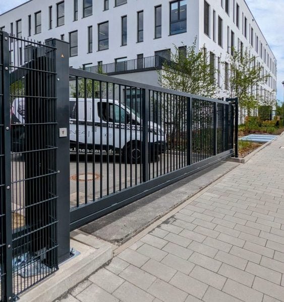 Moderne weiße Mehrfamilienhaus mit mehreren Fenstern, umgeben von jungen Bäumen, Parkplatz mit weißen Lieferwagen, umgeben von schwarzem Metallzaun, gepflasterter Gehweg im Vordergrund.