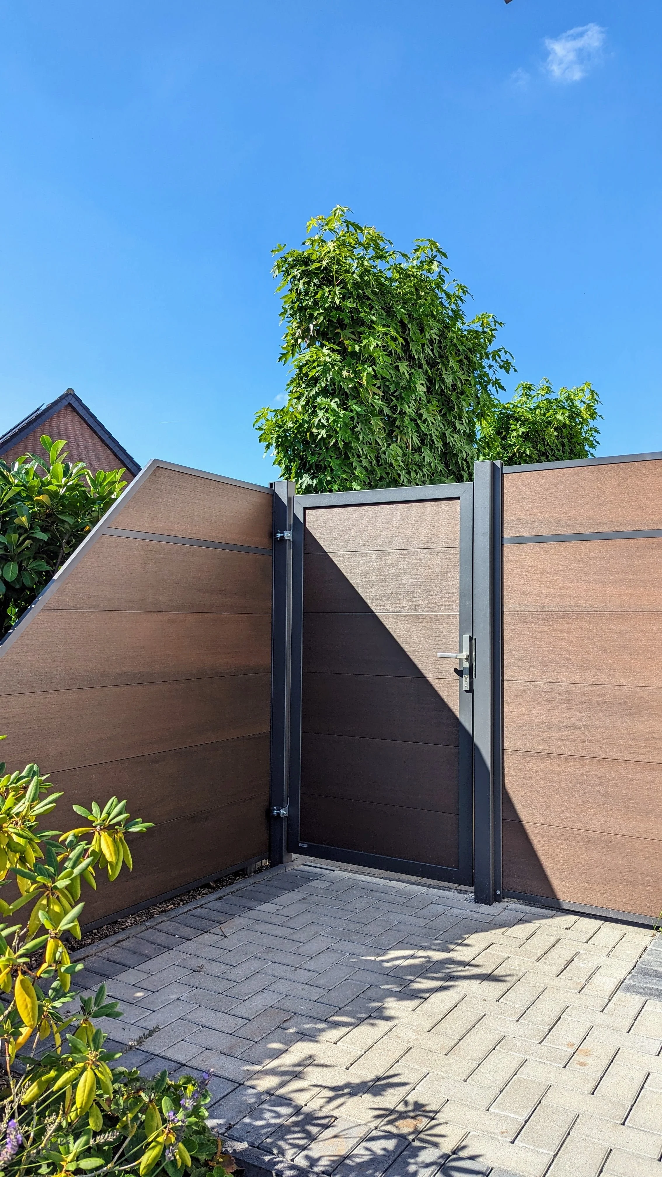 Hofeingang mit Holzzaun, grünen Pflanzen und Baum, blauer Himmel