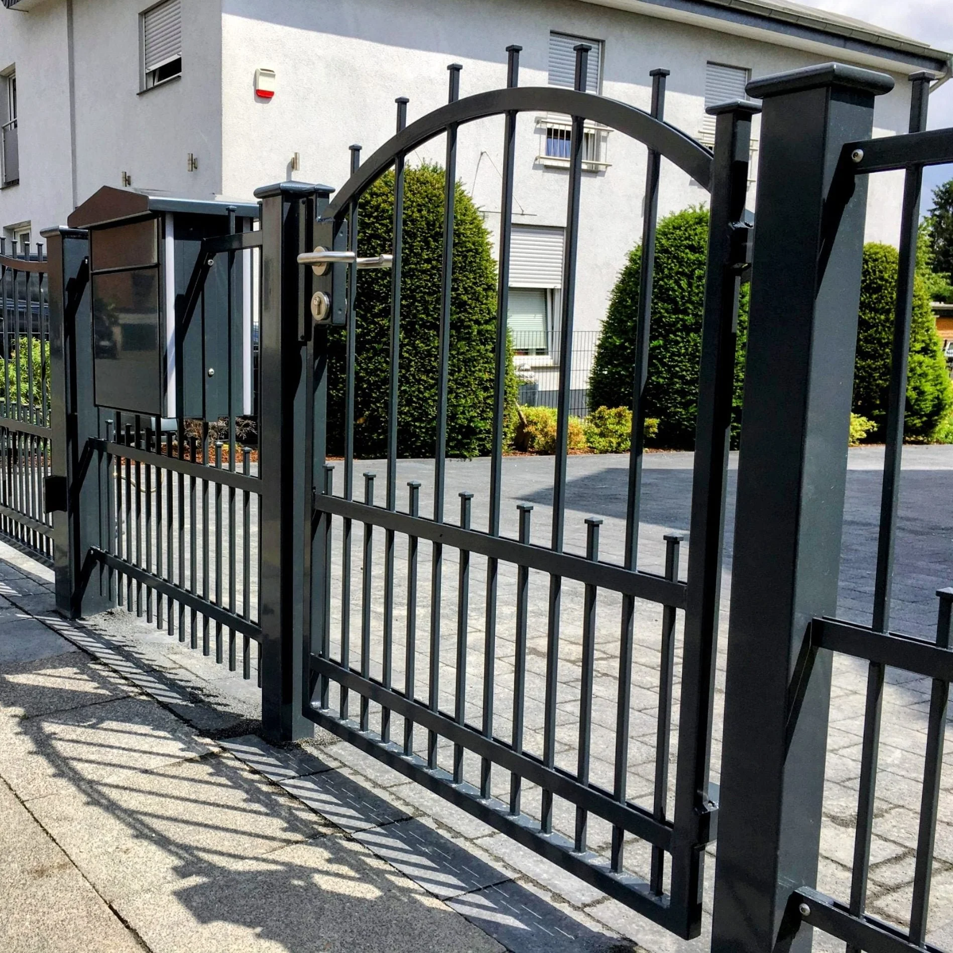 Schwarzes Metalltor mit Zaun vor mehrstöckigem Wohnhaus und Büschen im Hintergrund.