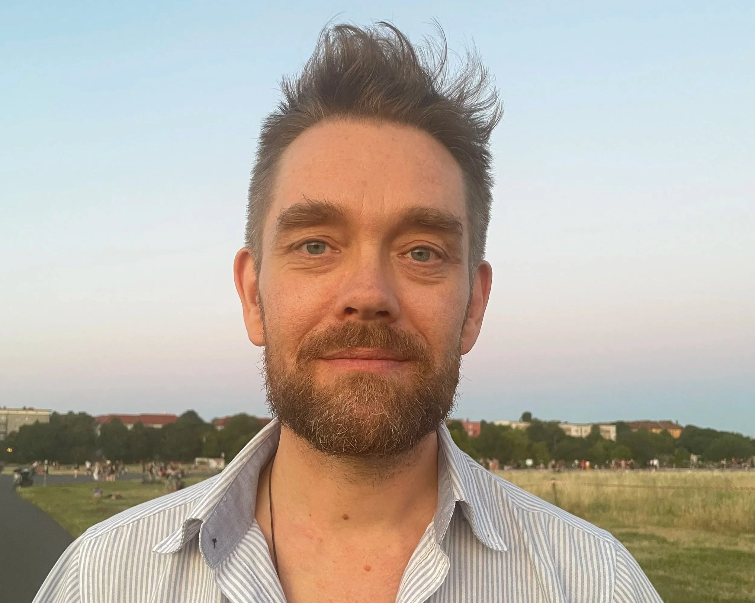 A man with a beard and mustache taking a selfie outdoors during sunset, with a park and buildings in the background.