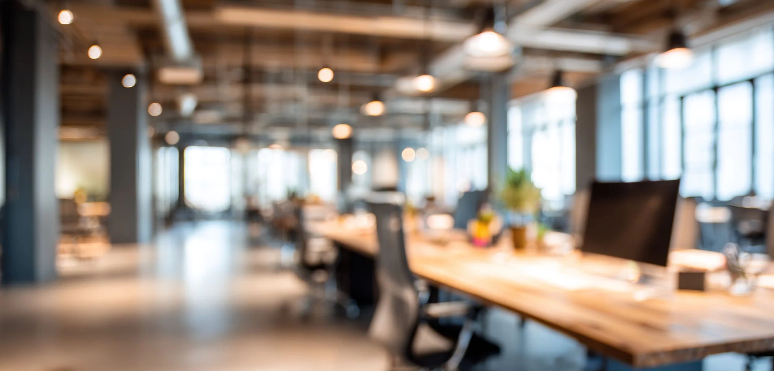 Blurred modern office space with large windows, wooden desks, black chairs, and computer monitors.