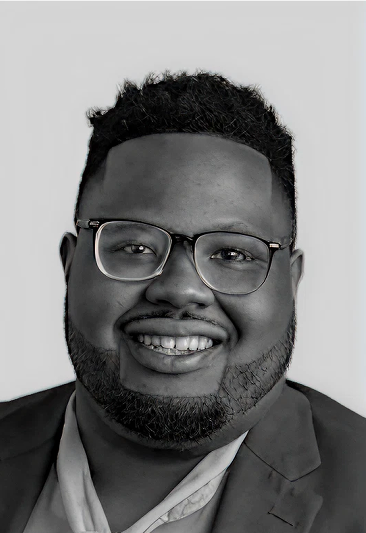A smiling man with glasses, a beard, and short curly hair, wearing a suit and tie, posed against a plain background.