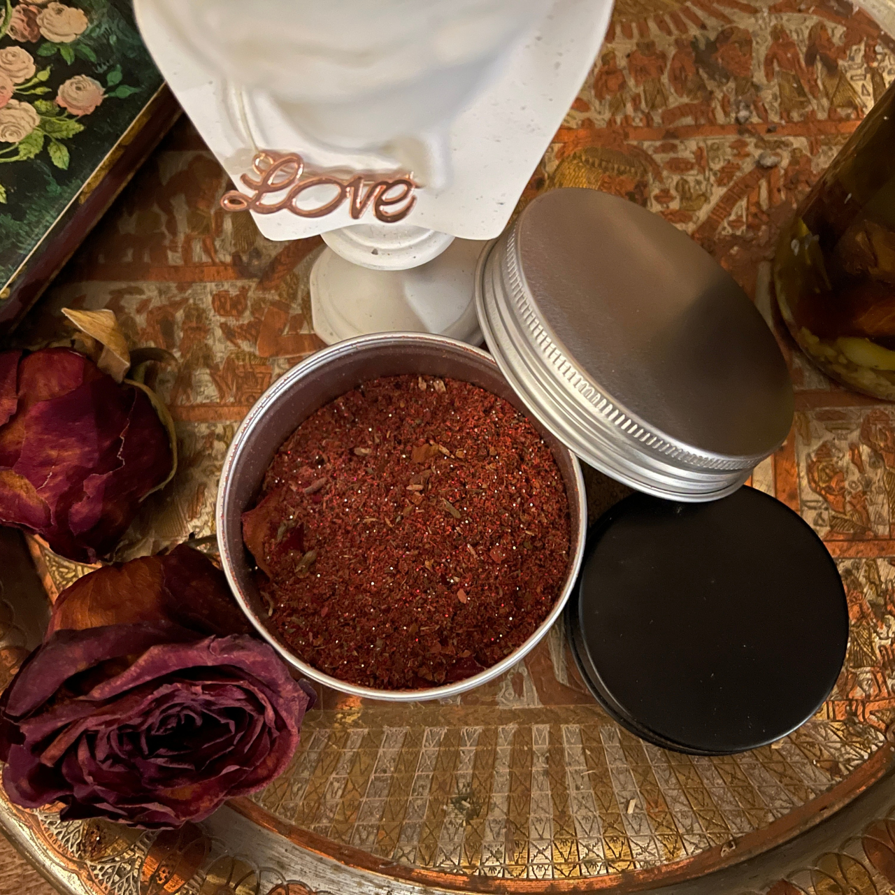 Open metal tins with dried herbs and spices, dried roses, and a decorative white container with a rose gold 'Love' sign on a decorative, textured surface.