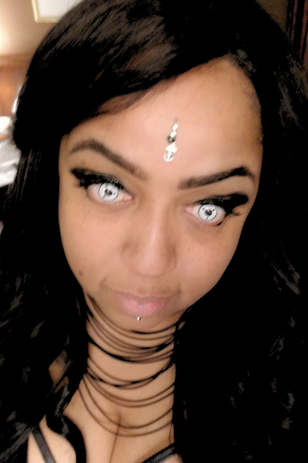 A woman with dark hair, special effects contact lenses, and facial piercings, wearing black makeup and a black choker, with a decorative forehead jewelry piece.