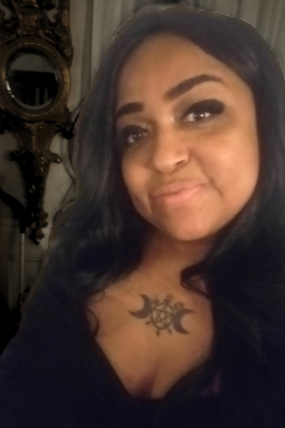 A woman with long dark hair, dark makeup, and tattoos on her chest, smiling slightly, in a dimly lit room.