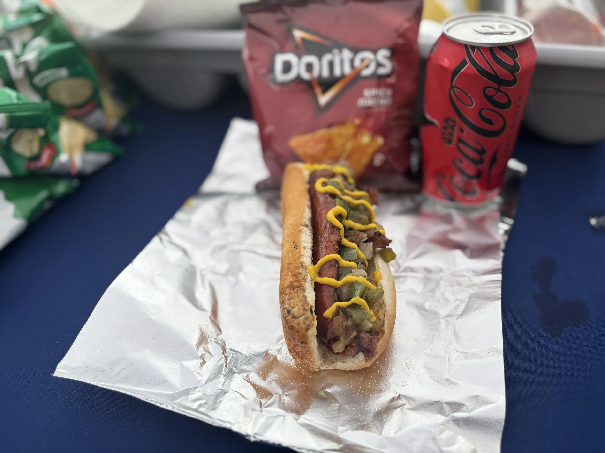 A hot dog with relish and mustard on a piece of foil with a bag of Doritos