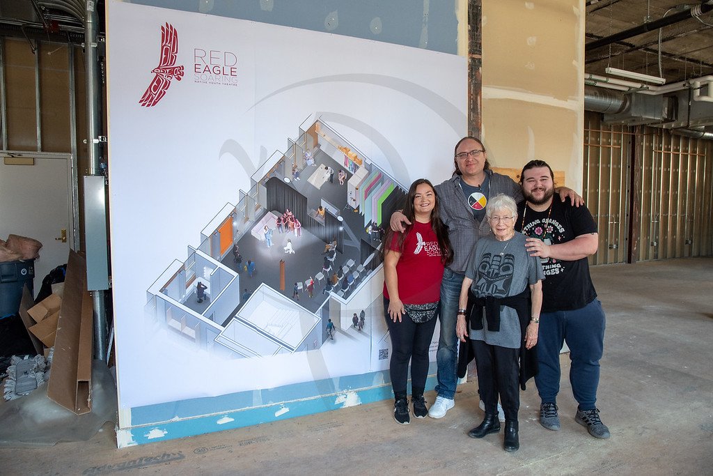 Four people standing in front of an architectural blueprint for the Red Eagle Sagoon building, inside a construction site.