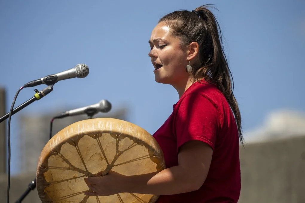 Young Native American woman singing into microphones, playing a drum outdoors on a sunny day.