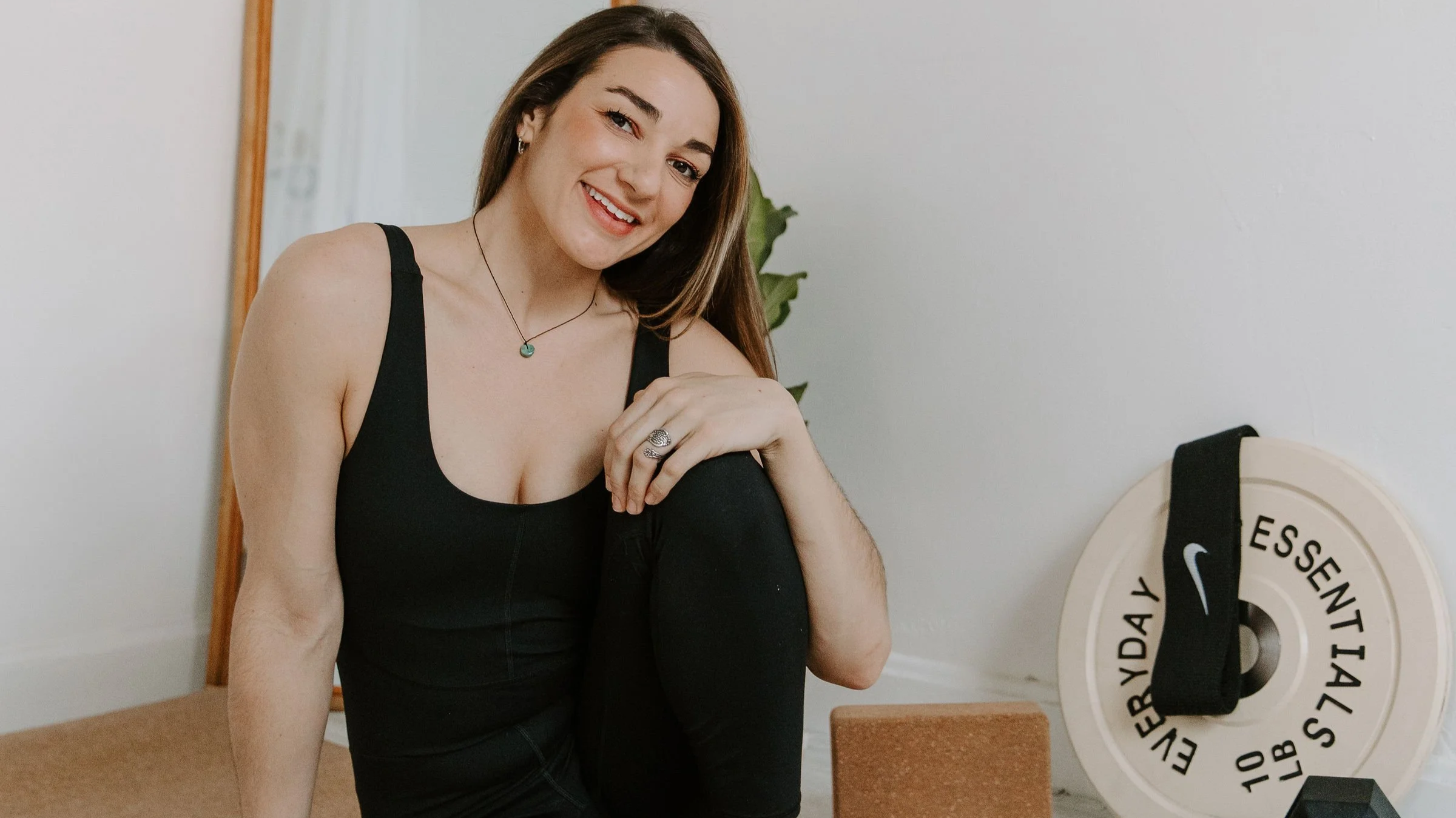 A woman in workout attire sitting on the floor in a room with exercise equipment and a mirror.