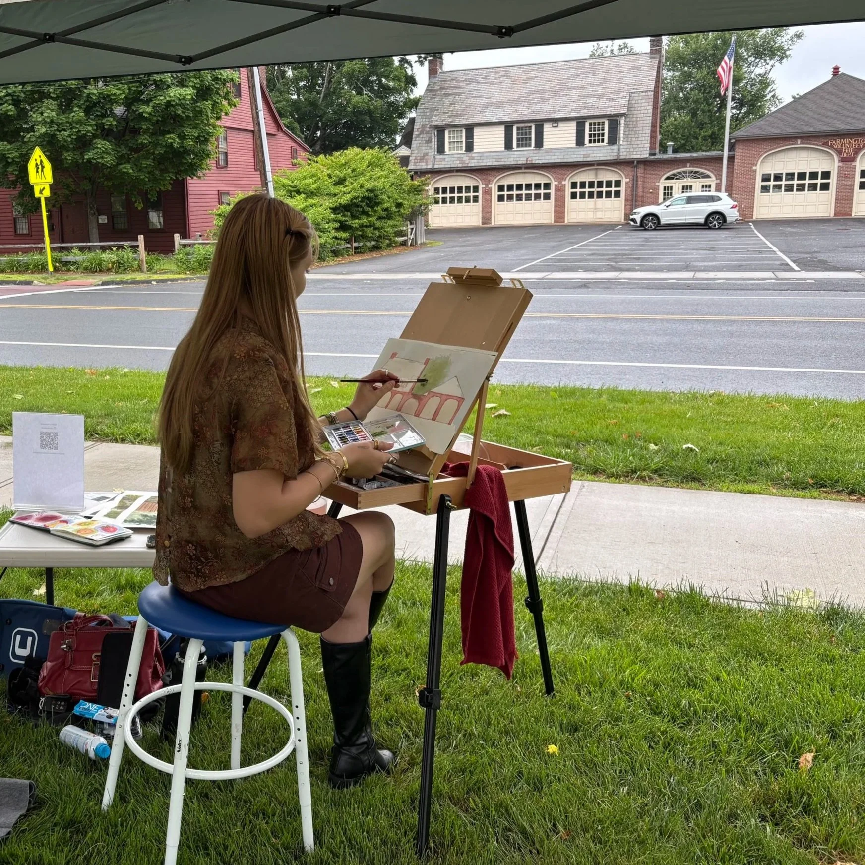 A woman painting a landscape on an easel outdoors under a tent, with a street, parking lot, and brick and wooden houses in the background.