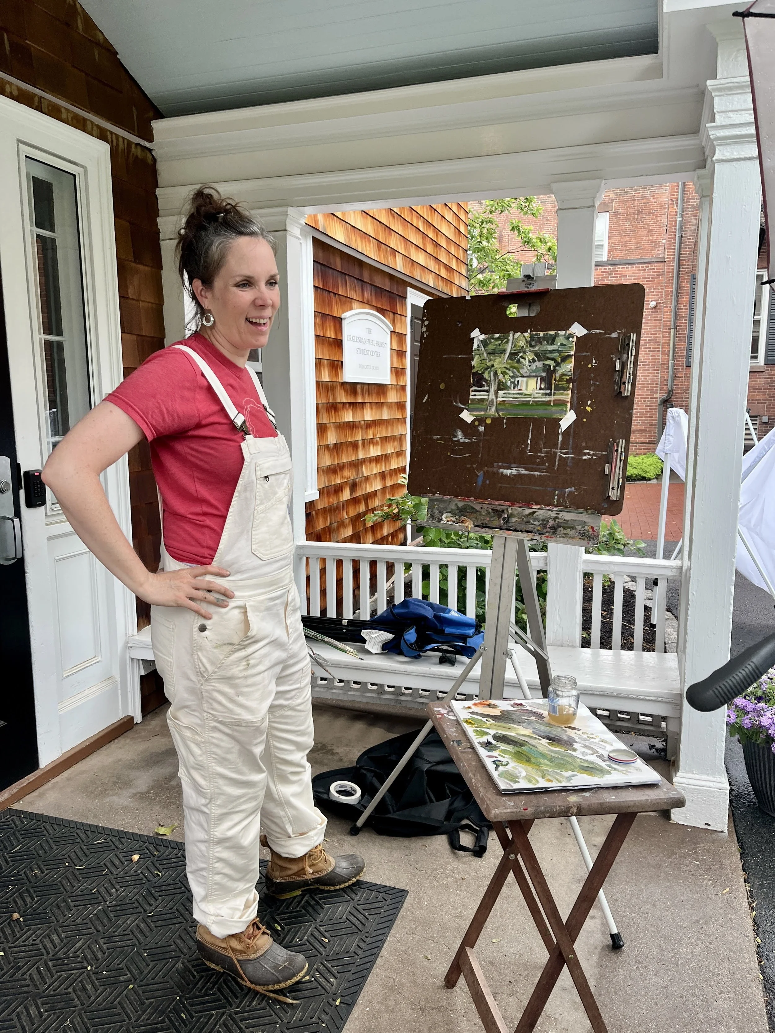 Woman in a red shirt and white overalls standing on a porch next to an easel with a mirror, painting supplies on a small wooden table, and a porch bench with a bag. She is smiling and looking at her painting.