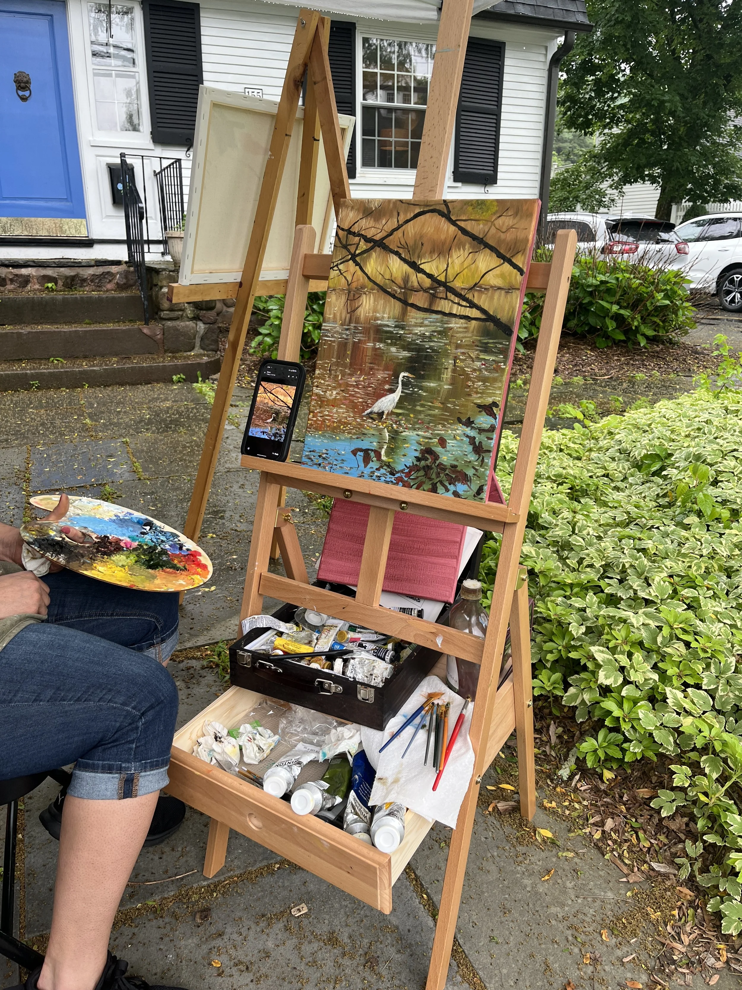 An outdoor artist's setup with a painting of a heron in a pond, a paint palette, brushes, and tubes of paint. The artist, partially visible, is sitting and holding a palette.
