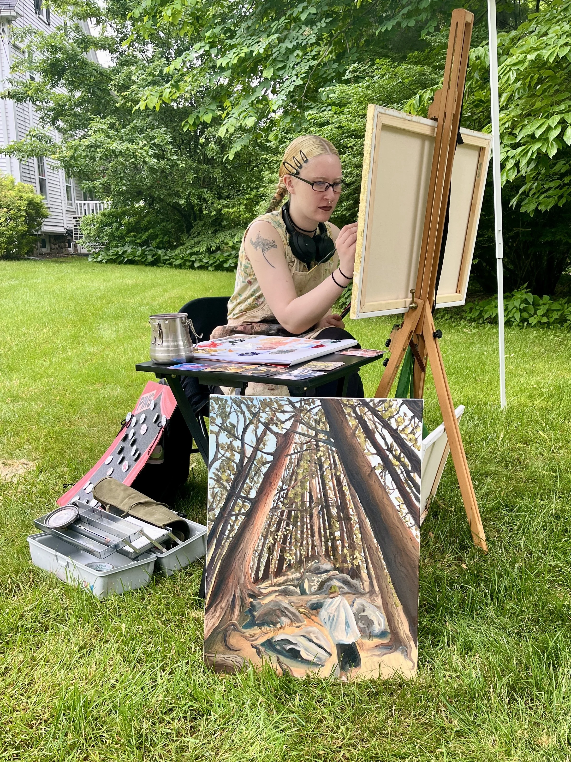 An artist paints outdoors in a park with trees and grass, sitting at an easel with a landscape painting of trees and rocks. Art supplies are on a small table and in a toolbox on the ground.
