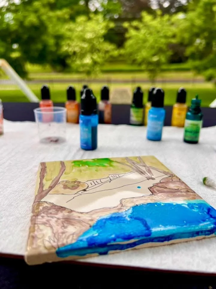 Watercolor painting of a landscape on a table with paint bottles in the background, set outdoors with green trees.