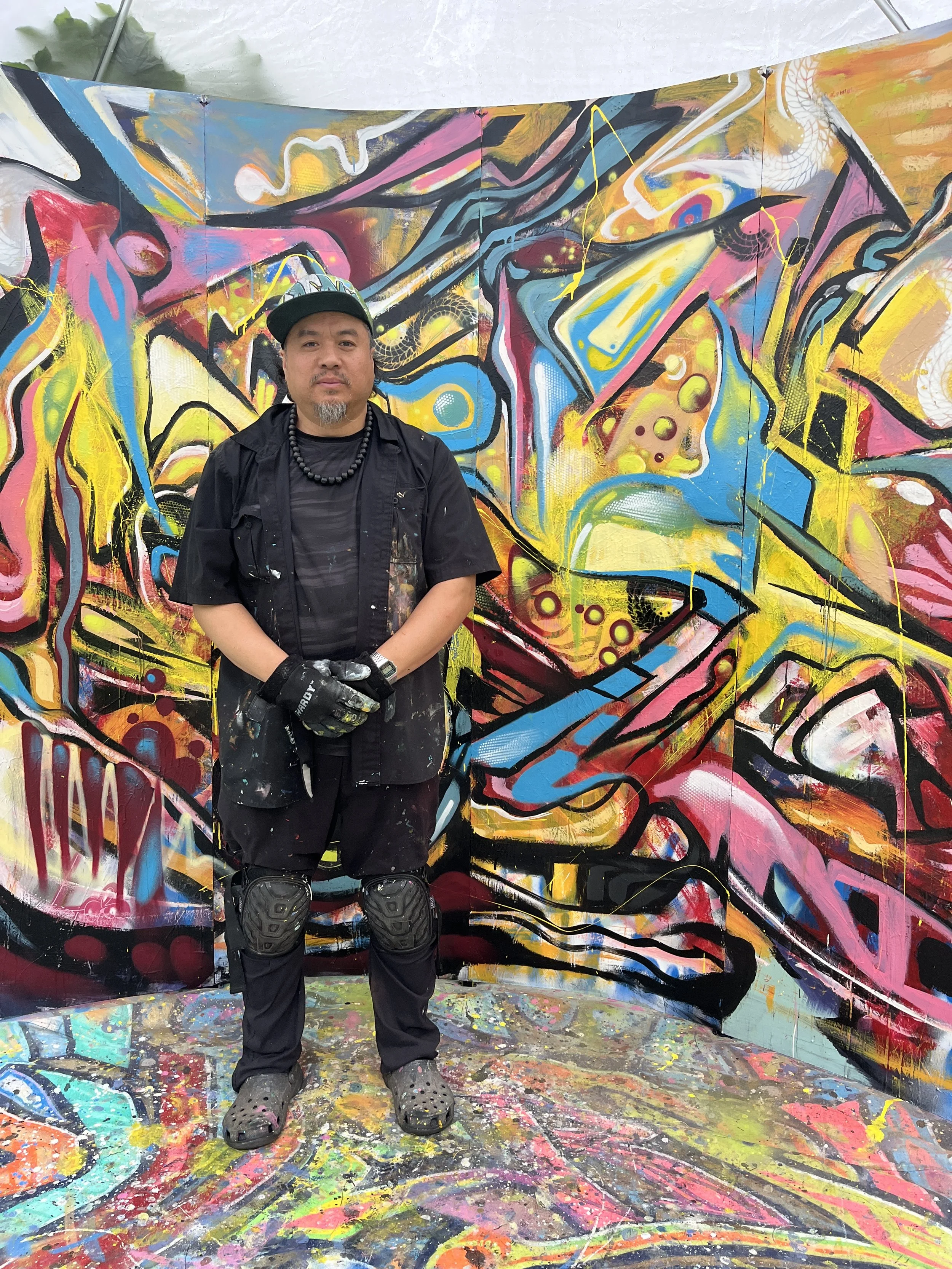 An artist in black clothing and graffiti gloves standing in front of a colorful, abstract graffiti mural with various bright colors and shapes.