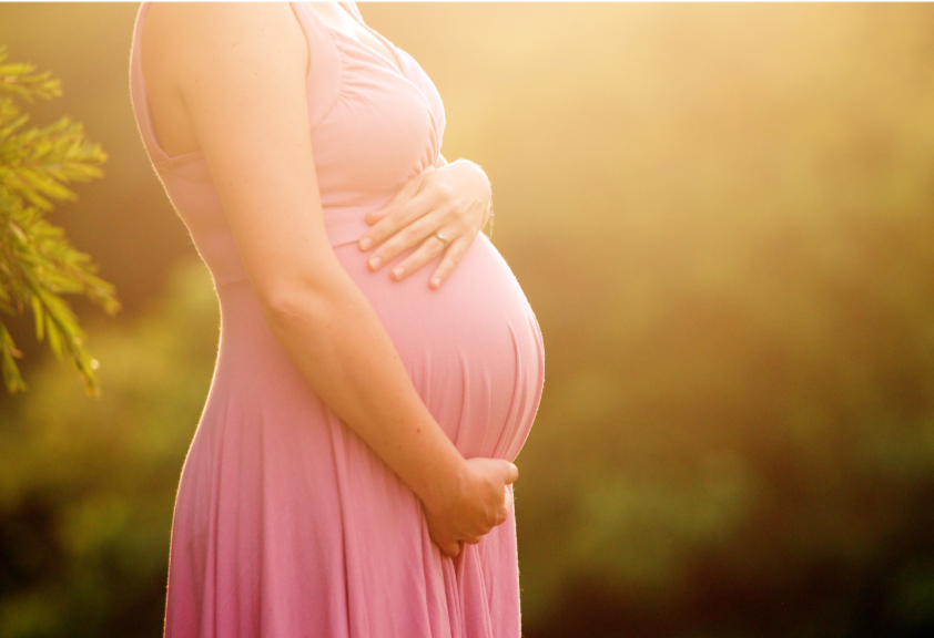 A pregnant woman in a pink dress holding her belly outdoors at sunset or sunrise.
