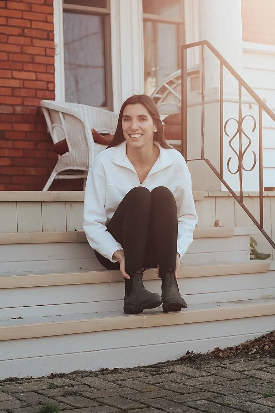 Young woman sitting on front porch steps, smiling, wearing a white jacket, black pants, and black boots, with a house and brick wall in the background.