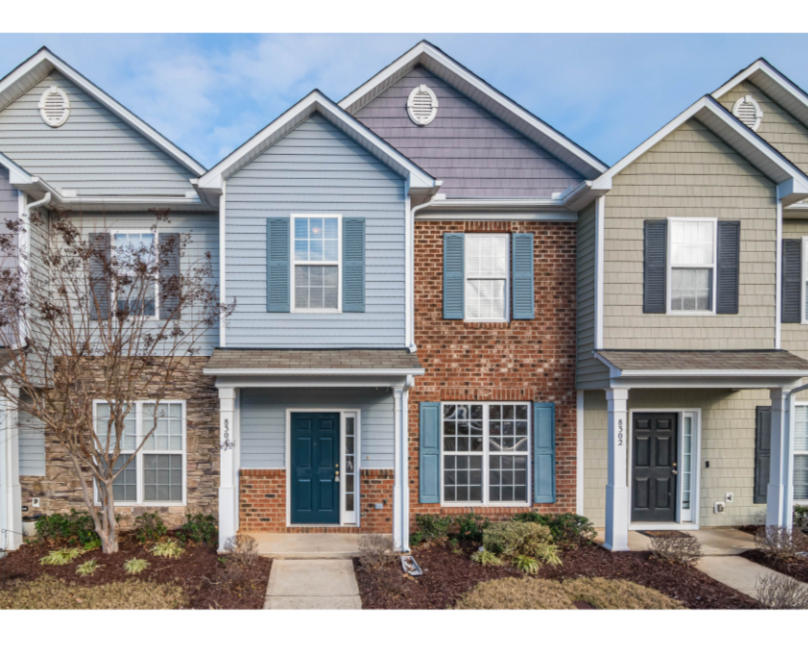 Row of townhouses with different siding colors, brick accents, and front doors, with small landscaped front yards and a cloudy sky.
