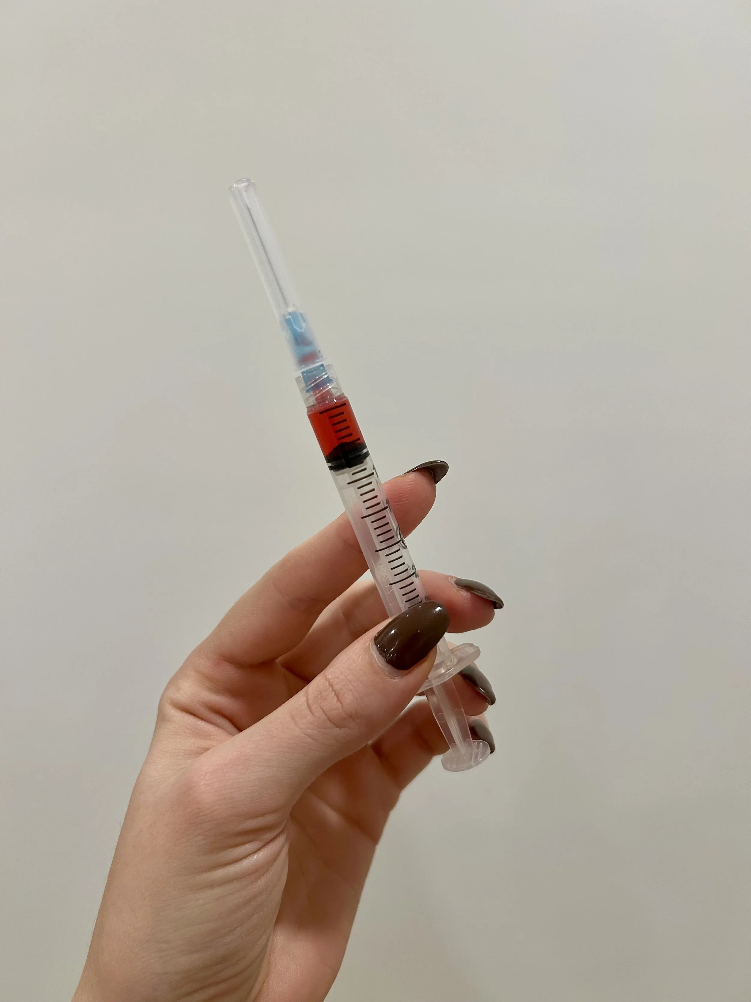 Close-up of a hand holding a syringe filled with red liquid against a plain background.