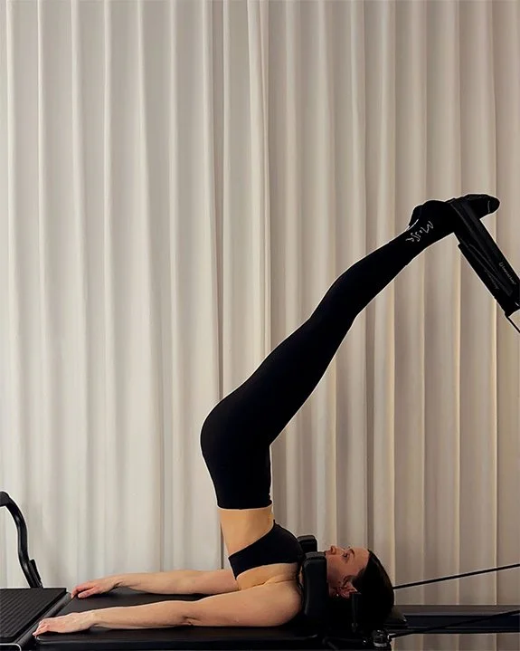 The Short Spine Pilates exercise on balanced body allegro reformer, inside a room with cream-colored curtains.