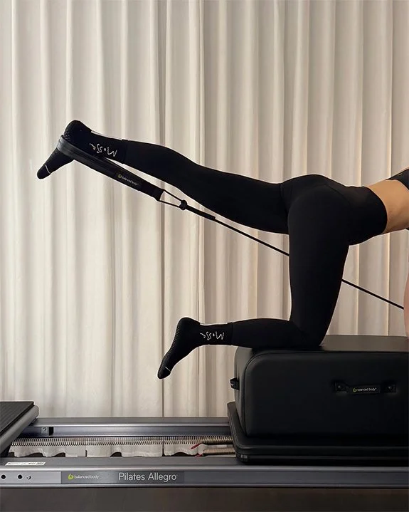 Person performing feet in strap kneeling on long box. Pilates exercise on a reformer machine, with one leg extended and wearing black leggings and socks.