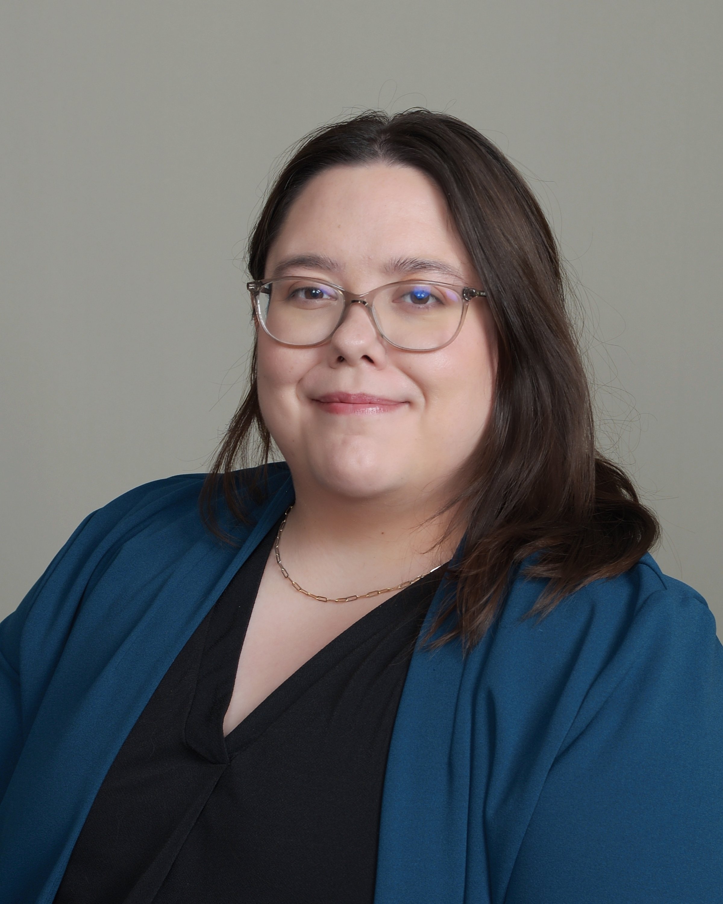 A woman with dark brown hair, wearing glasses, a black top, and a blue blazer, smiling against a plain background.
