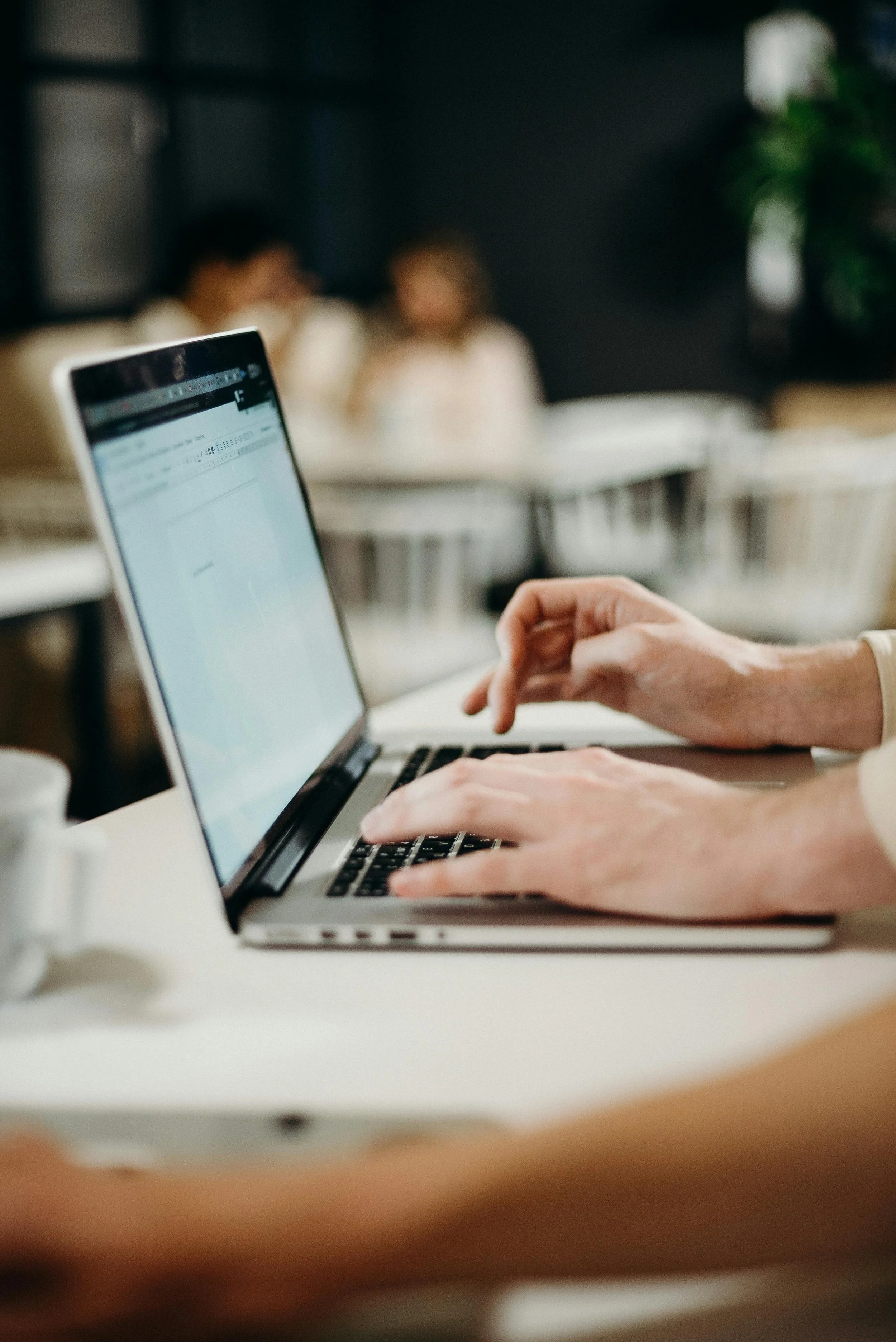 Person typing on a laptop and reporting a phishing email with two people blurred in the background at a table.
