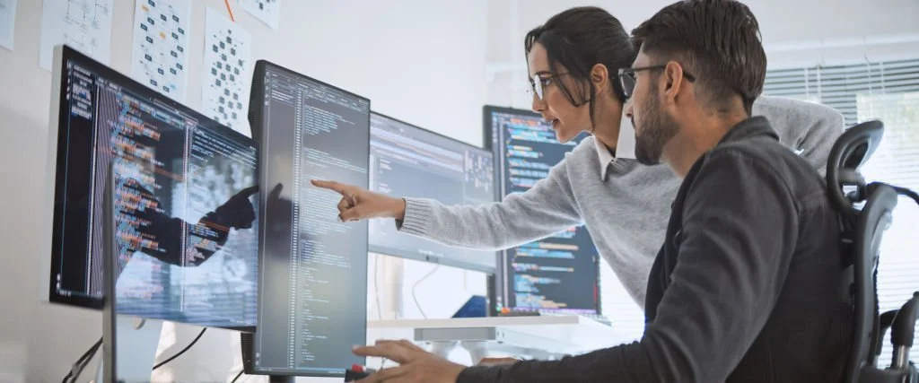 Two IT managers working together on a cybersecurity incident response plan at a desk with multiple computer screens displaying code, in a well-lit office.