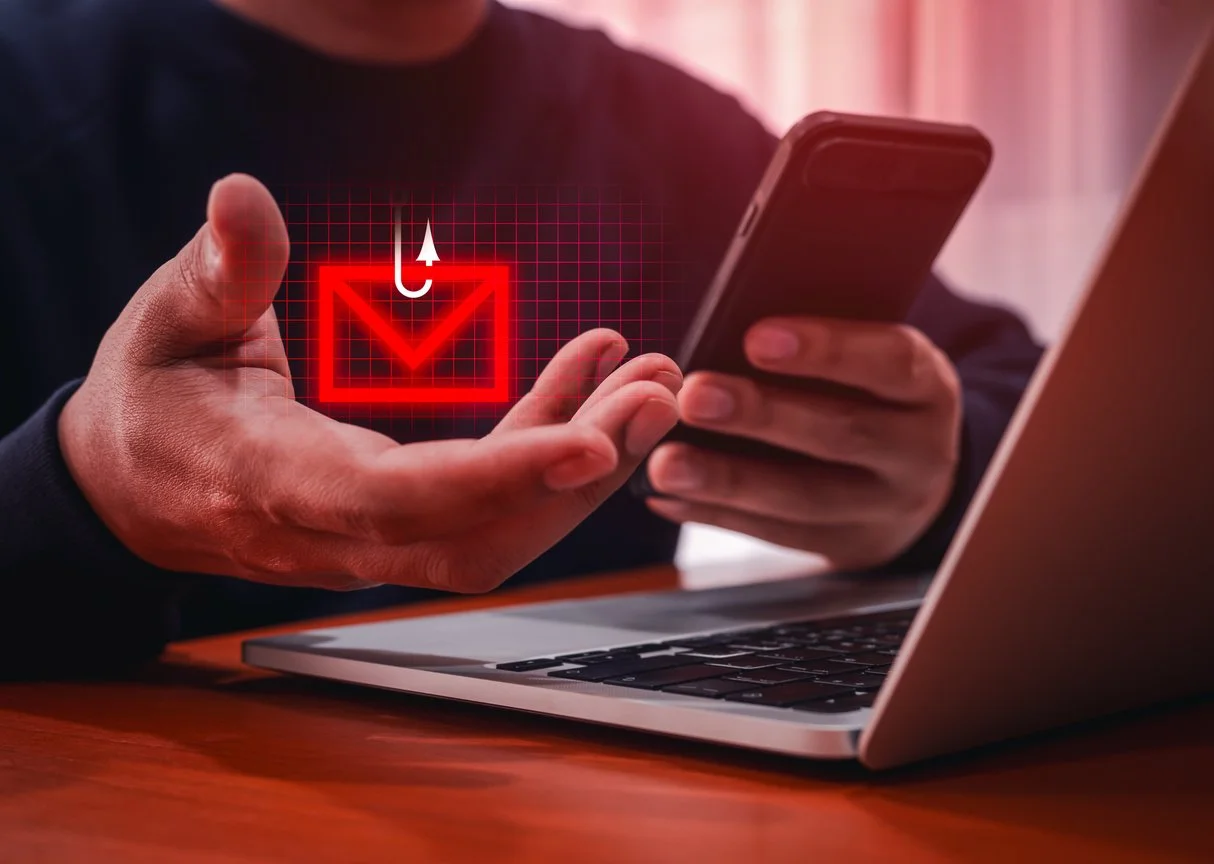 A person holds a smart phone in front of a laptop, a red email icon appears above his other hand with a fish hook through it.