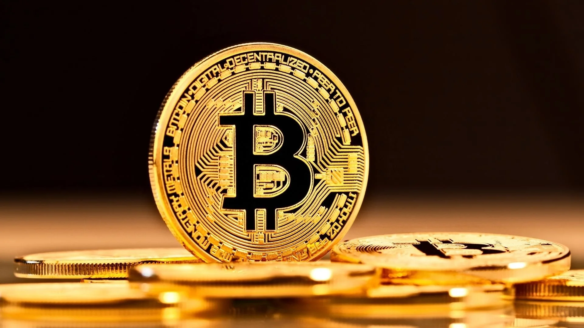 A shiny gold coin with a bit coin logo standing upright on other coins