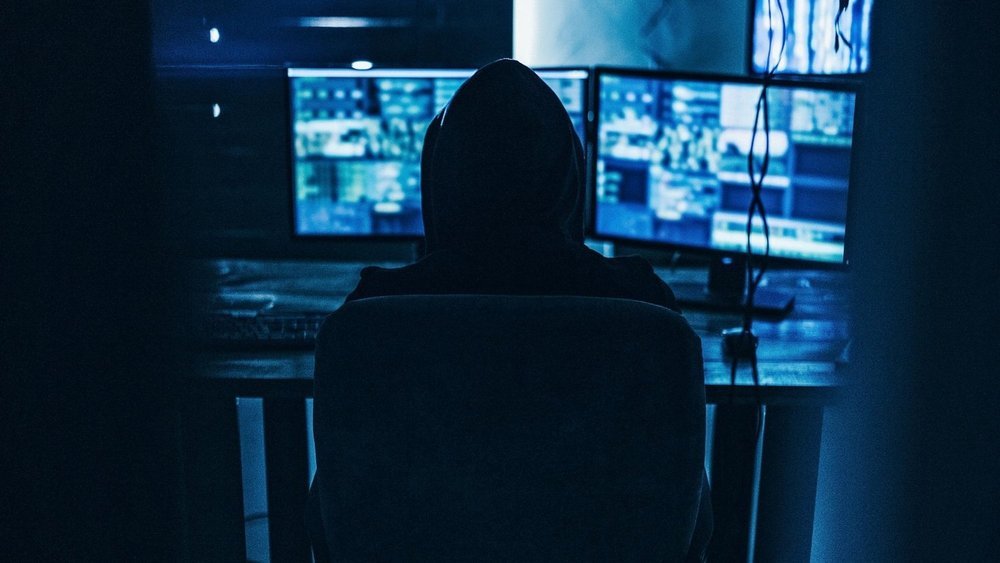 Picture of a hooded figure in the dark looking at computer screens.